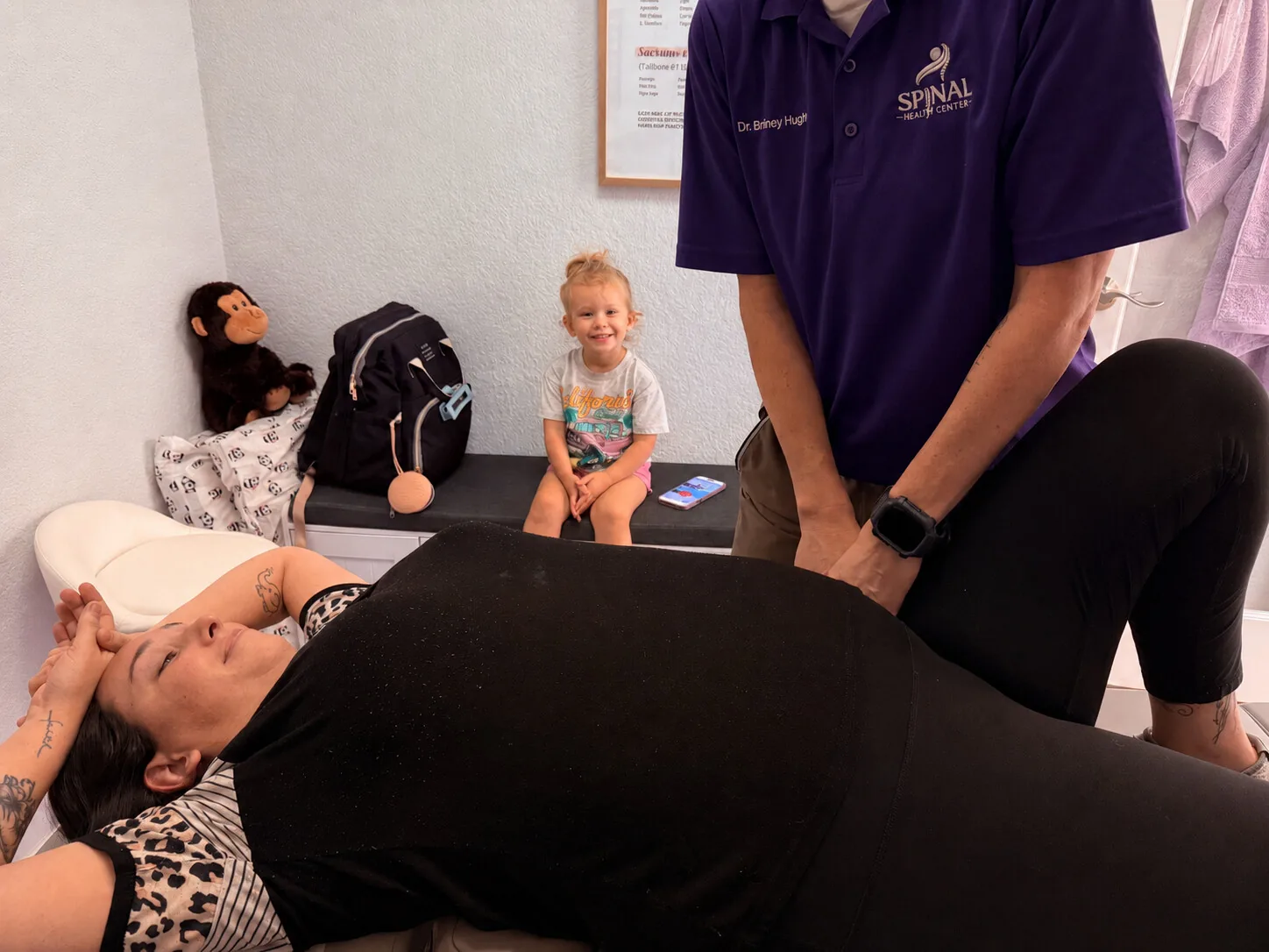 Pregnant patient receiving chiropractic care on a treatment table while a toddler looks on