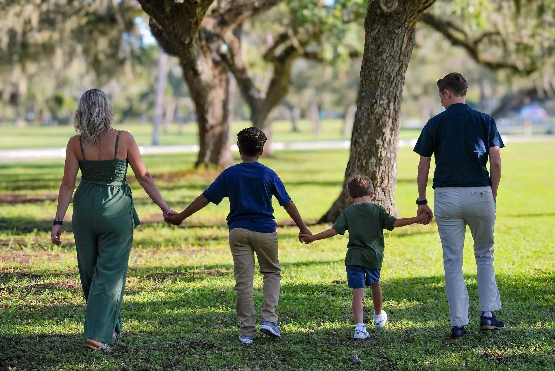 Family walking hand in hand across a park lawn under oak trees